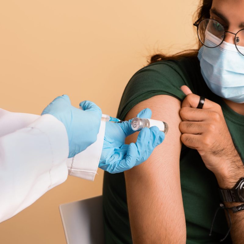 Closeup of nurse hands holding vaccine and syringe Closeup of nurse hands in medical gloves holding vaccine and syringe, doctor making vaccination against COVID-19 for middle-eastern millennial guy wearing protective mask, beige studio background
